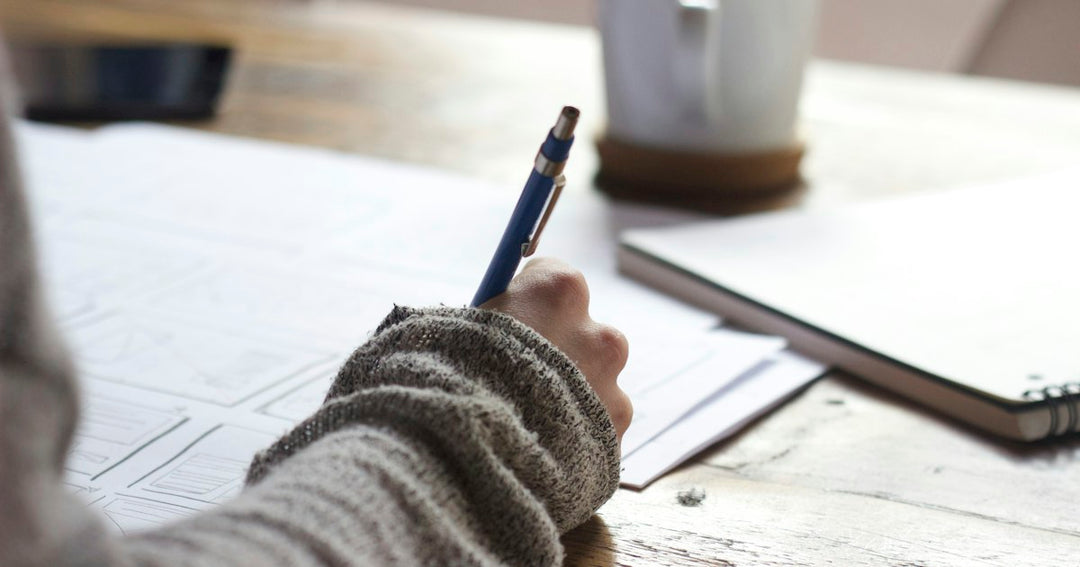 Une main tient un stylo bleu au-dessus de feuilles posées sur un bureau en bois clair, comme pendant l’épreuve d’expression écrite du TCF