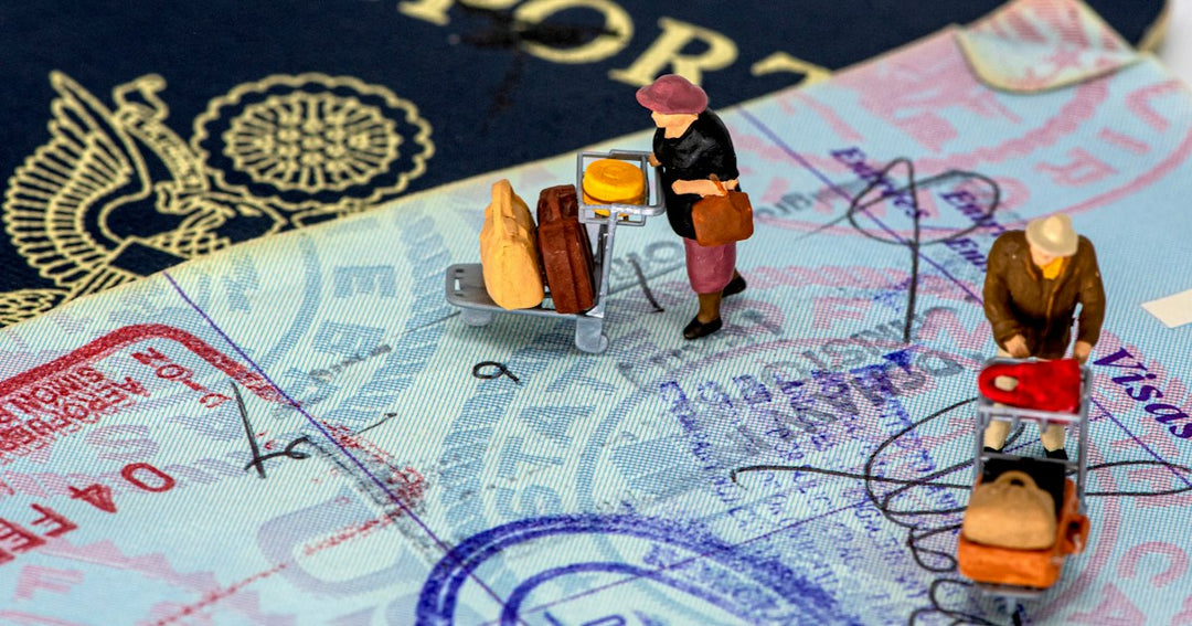 Deux figurines miniatures avec des chariots à bagages posées sur une page de passeport couverte de tampons, image d’une démarche d’immigration